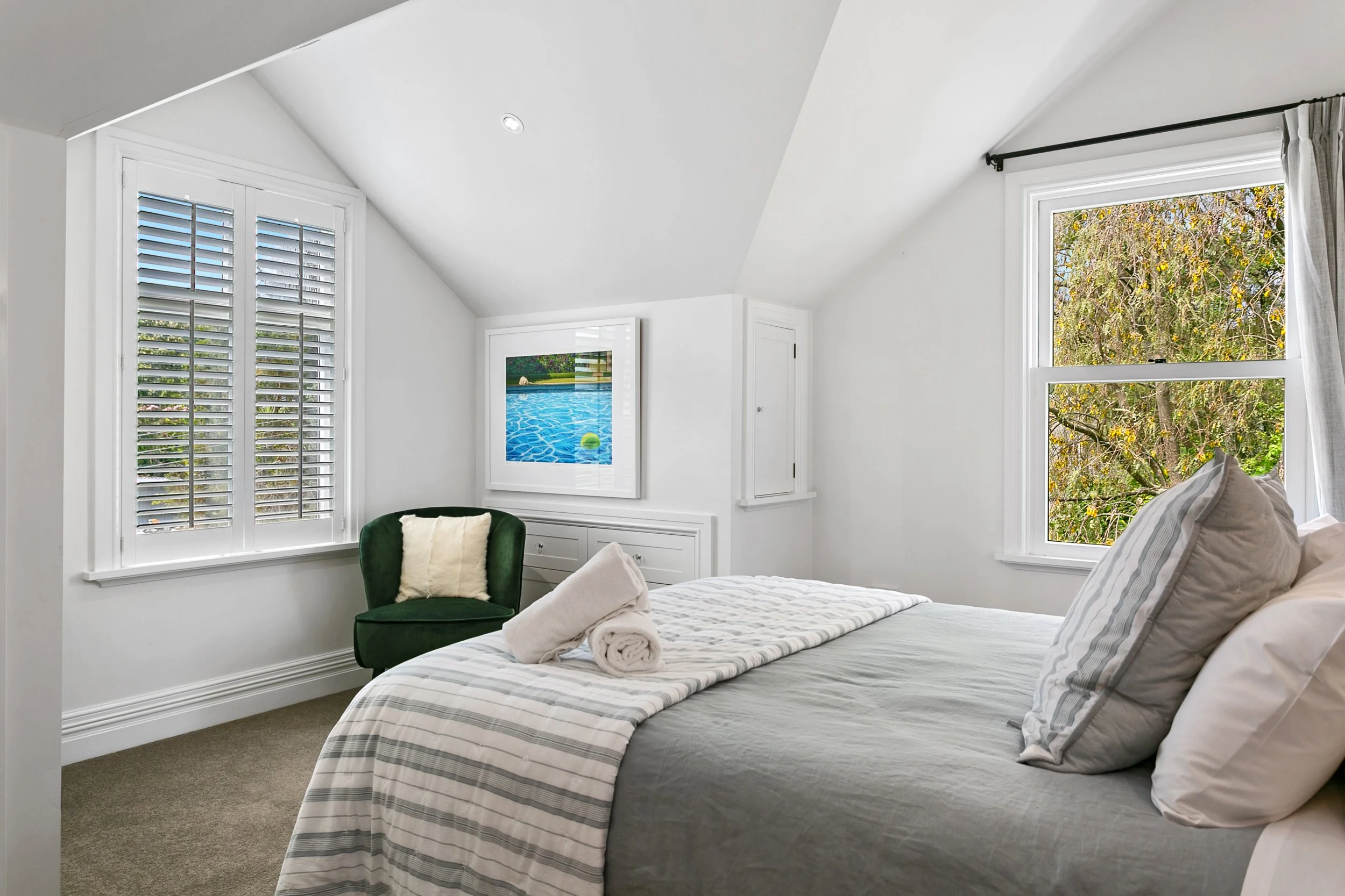 A neatly made bed with rolled towels on top, situated in a bright white bedroom with two large windows showing trees outside. There is a green armchair with a pillow, a framed picture of a swimming pool on the wall, and built-in white cabinets.