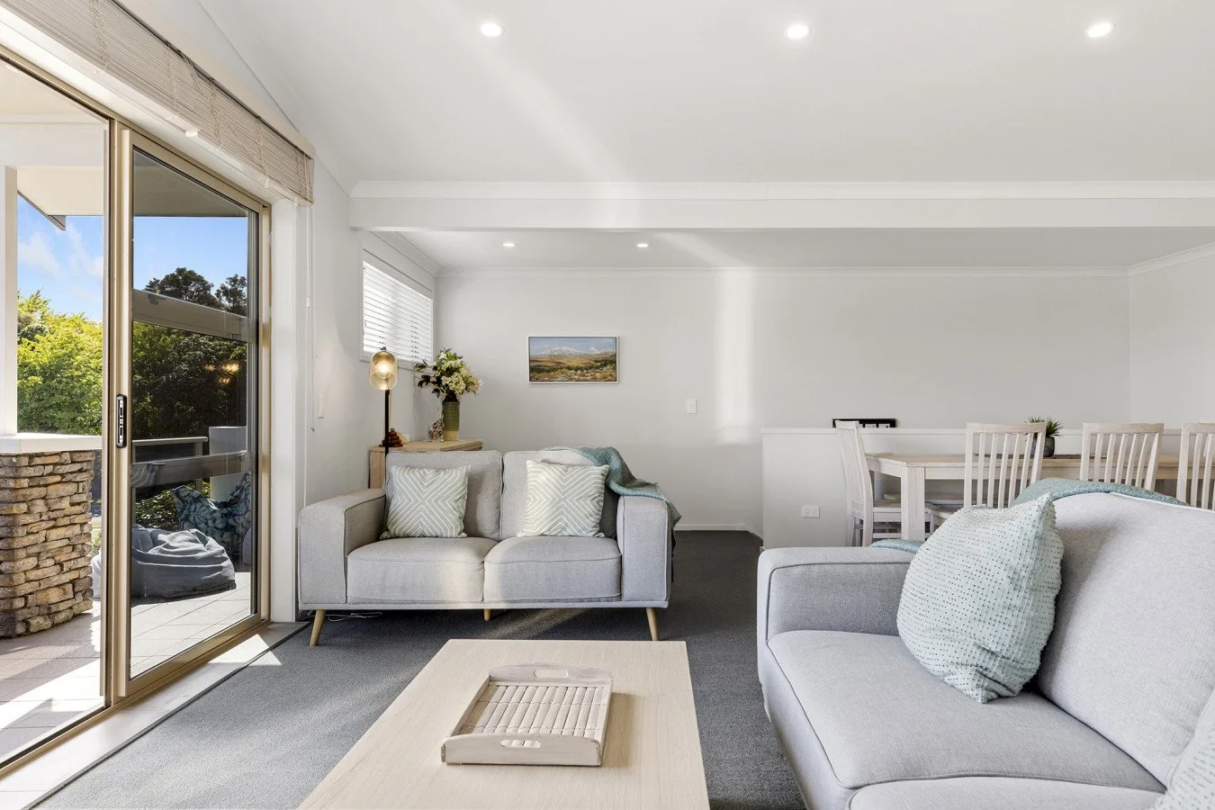 Bright living room with sliding glass door leading to balcony, white sofas with patterned pillows, wooden coffee table, and dining area with white table and chairs, decorated with artwork and plants.