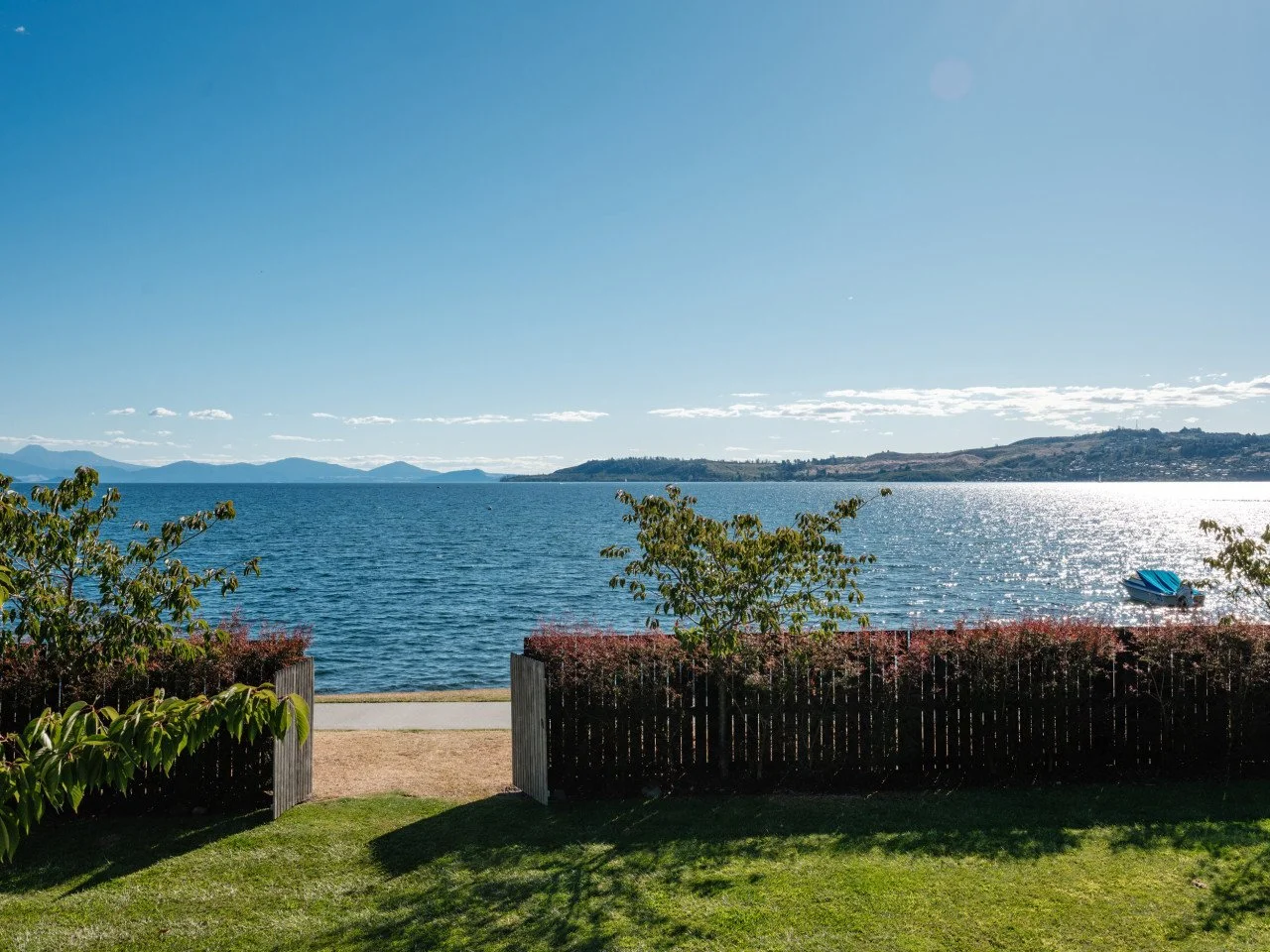 View of a large body of water with distant hills and partly cloudy sky, taken from a grassy yard with a wooden fence and small trees, and a boat near the shoreline.