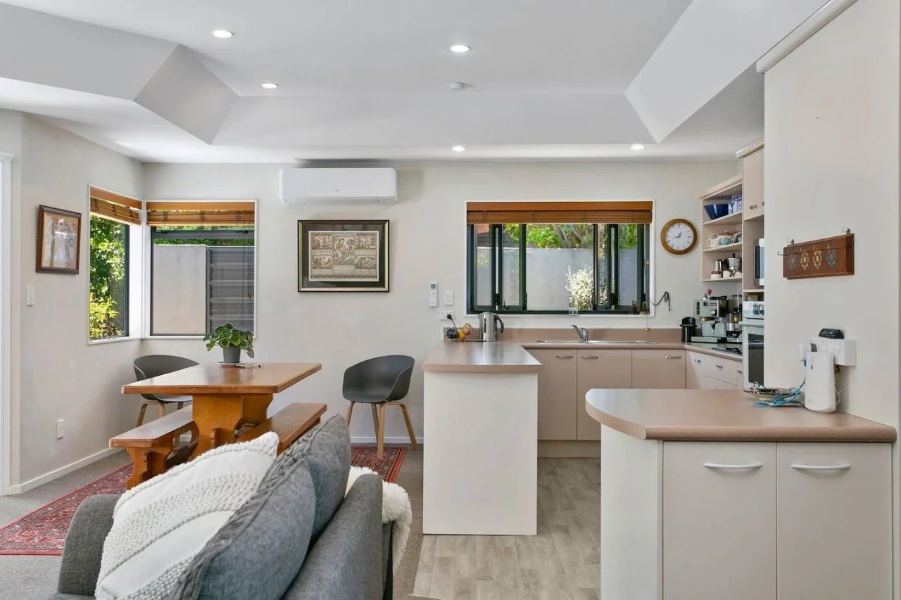 Open-concept kitchen and dining area with white walls, wooden cabinet countertops, and a window view of greenery. Features include a dining table with benches, a sofa with a knitted blanket, and kitchen appliances.