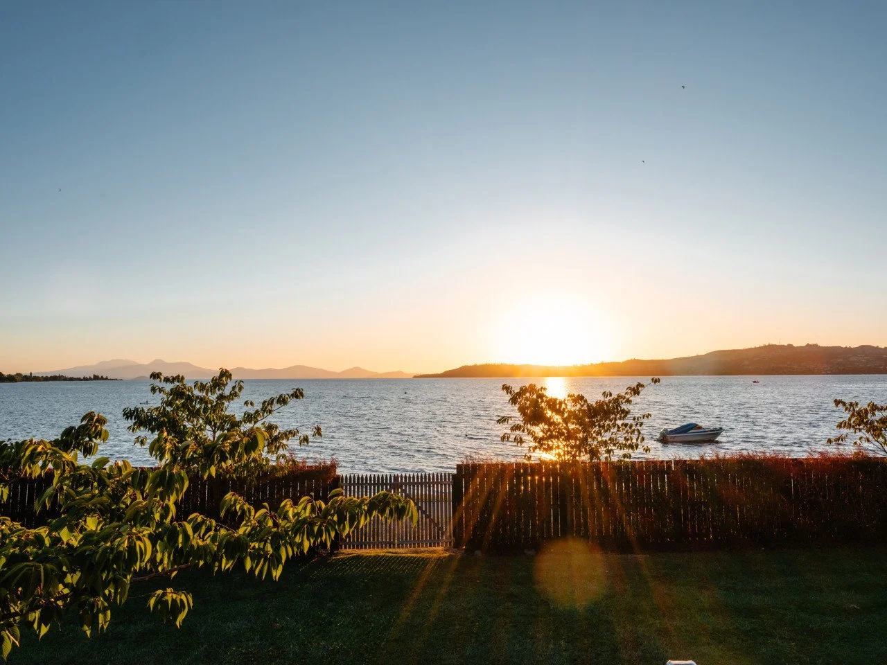 Sunset over a body of water with a small boat, trees, and a fence in the foreground.