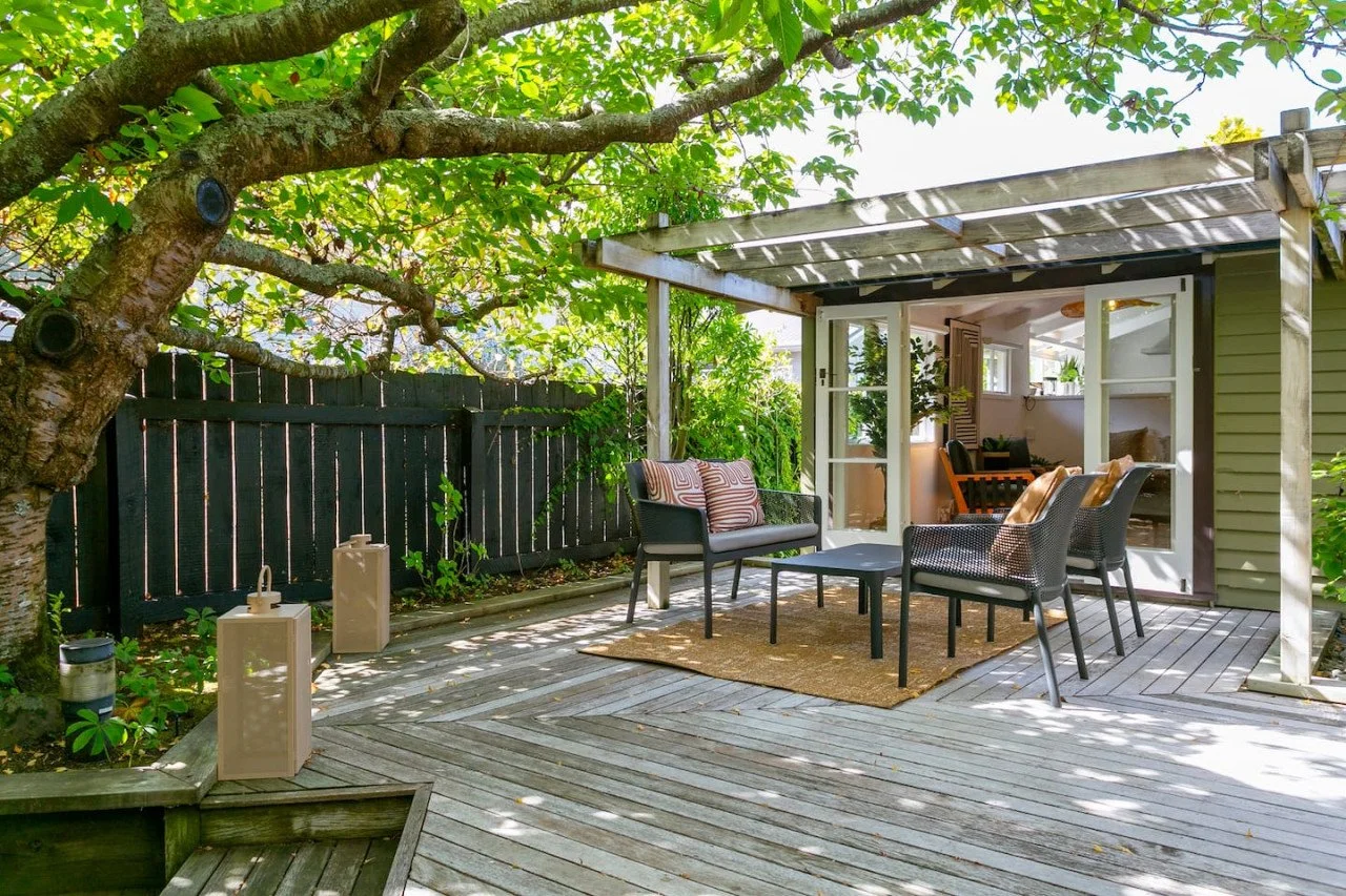 Outdoor patio with wooden deck, black fence, and greenery including a large tree. There is outdoor seating with a small table and decorative pillows. An open door reveals a cozy indoor space.