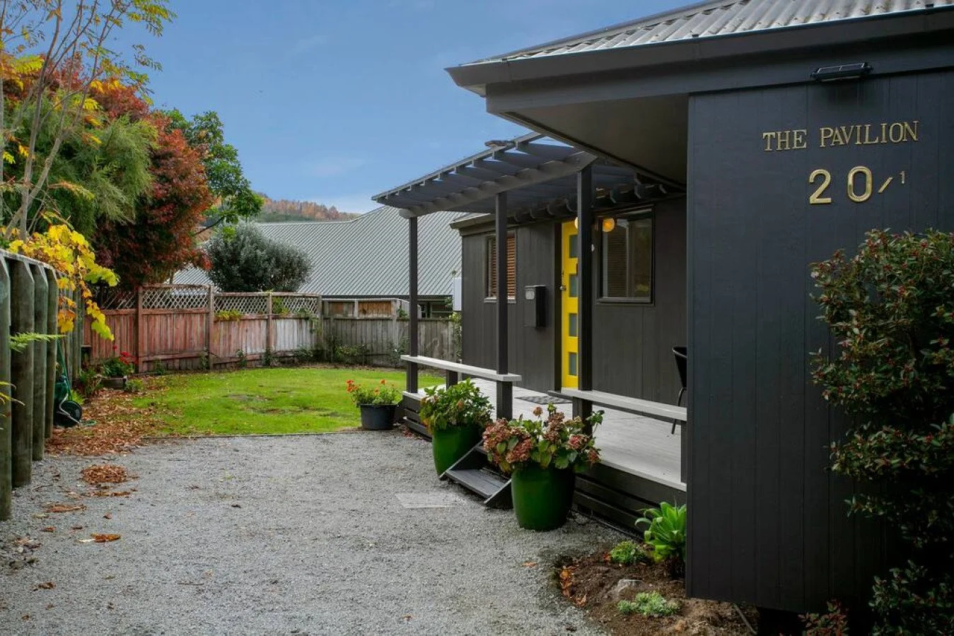 Front yard of a modern black house with a yellow door, potted plants, gravel driveway, and a wooden fence surrounded by trees.