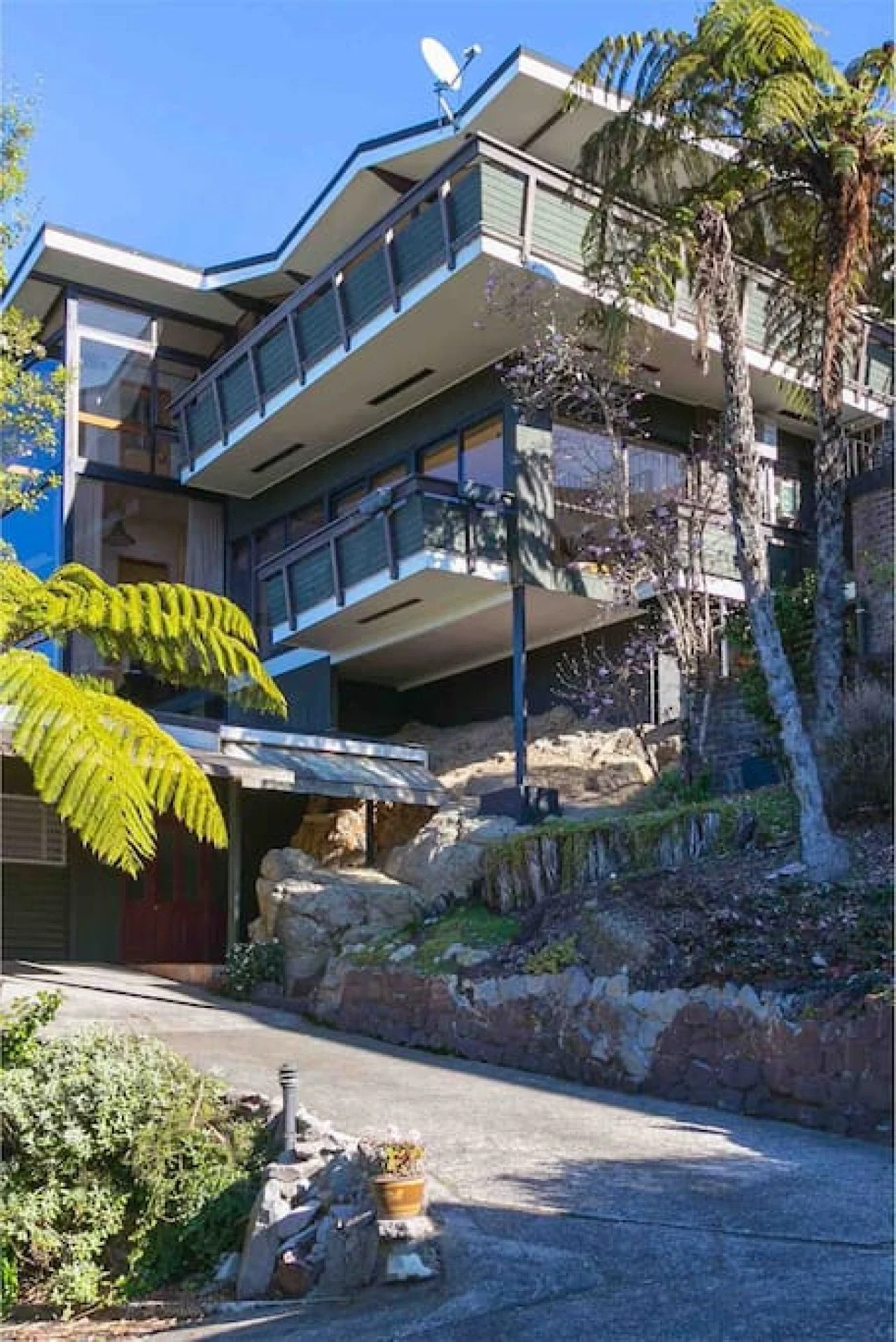 A multi-story modern house built on a sloped terrain, with balconies on each level surrounded by glass and metal railings, and lush trees and plants nearby.