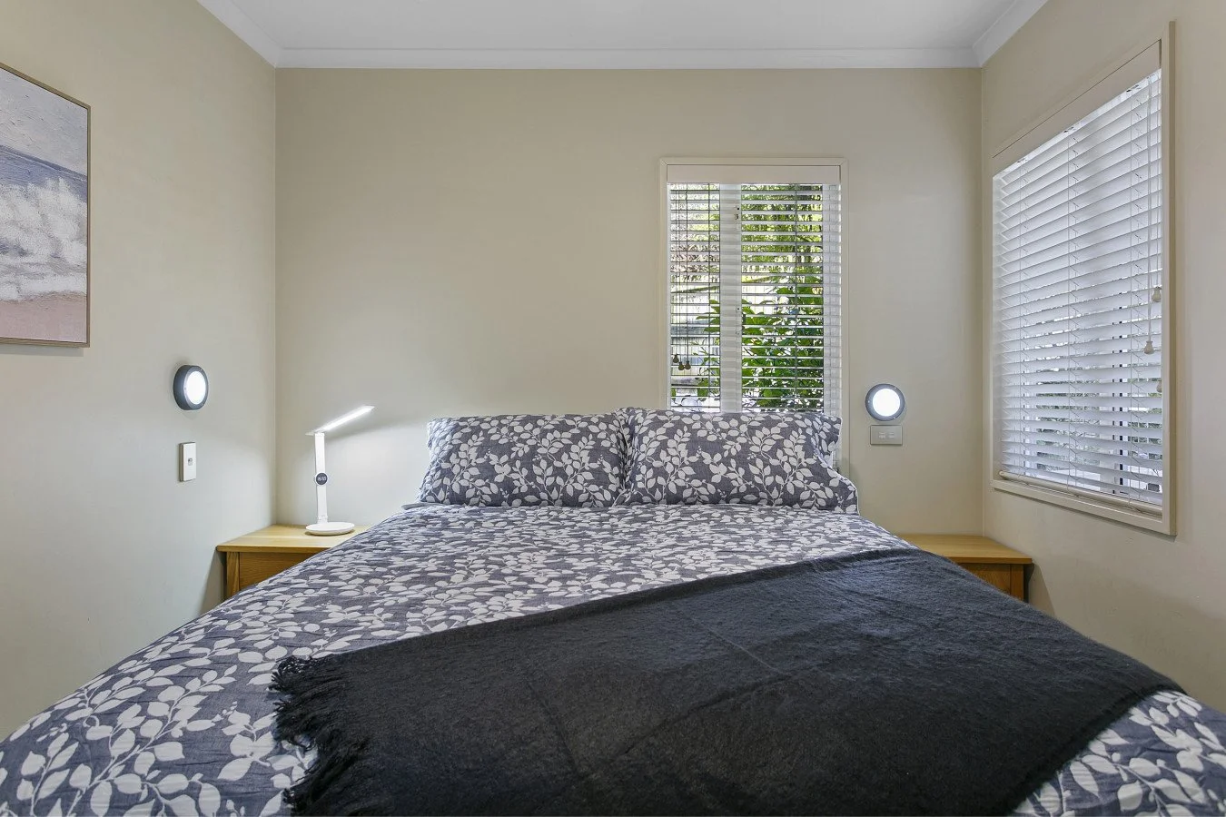 A neatly made bed with floral-patterned bedding and pillows, flanked by wooden nightstands with lamps, in a bedroom with cream walls and two windows with white blinds.