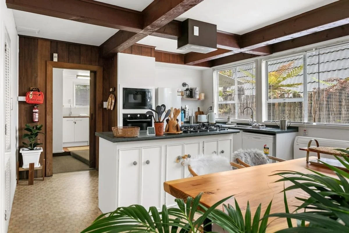A kitchen with white cabinets, a black countertop, and large windows with blinds. There are wooden beams on the ceiling, and a wooden dining table in the foreground. The kitchen has various small appliances, utensils, and plants, with a wood-paneled 