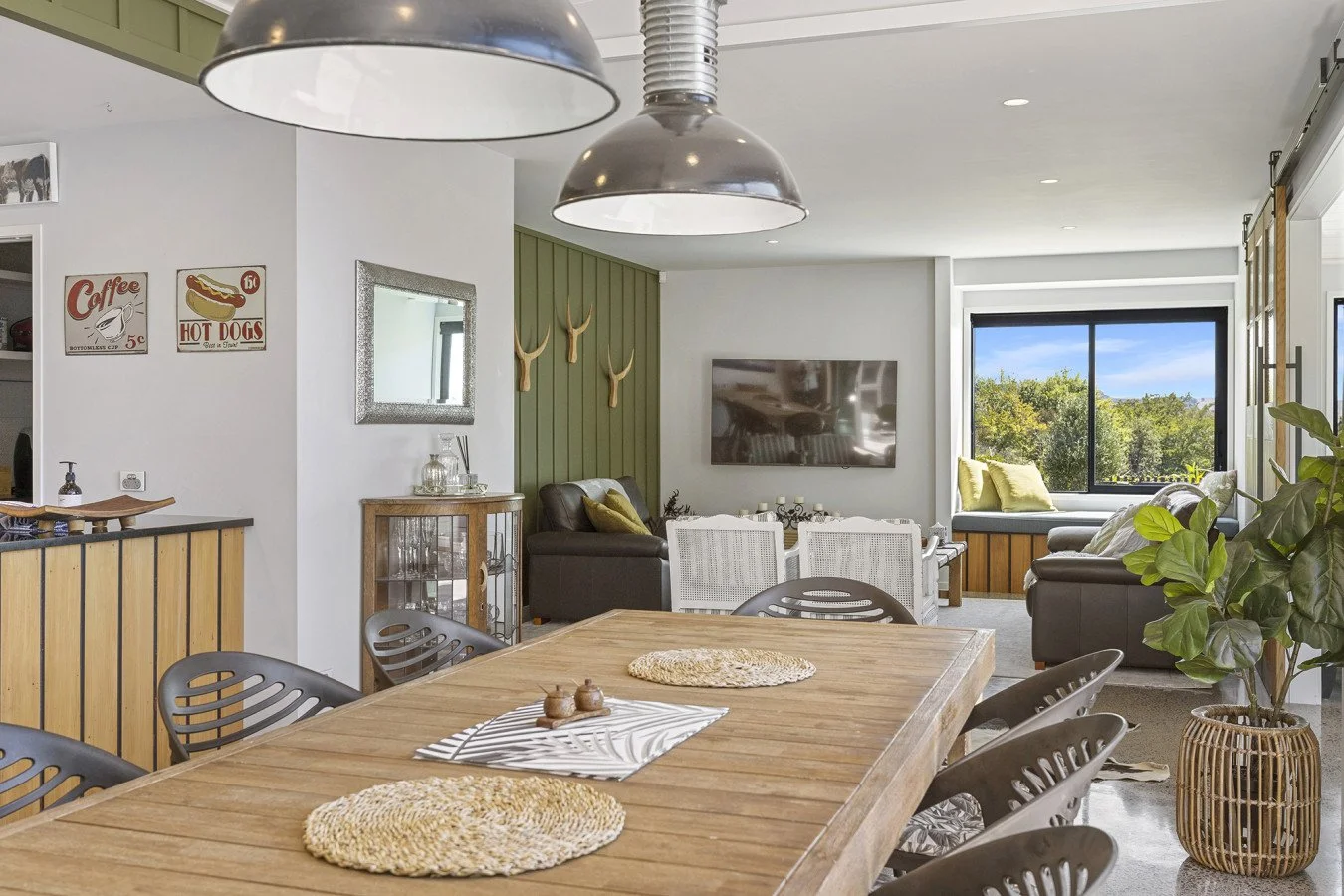 Open-concept living and dining area with a wooden dining table, black chairs, a potted plant, a green accent wall with deer antlers, a television, and a large window showing outdoor greenery.