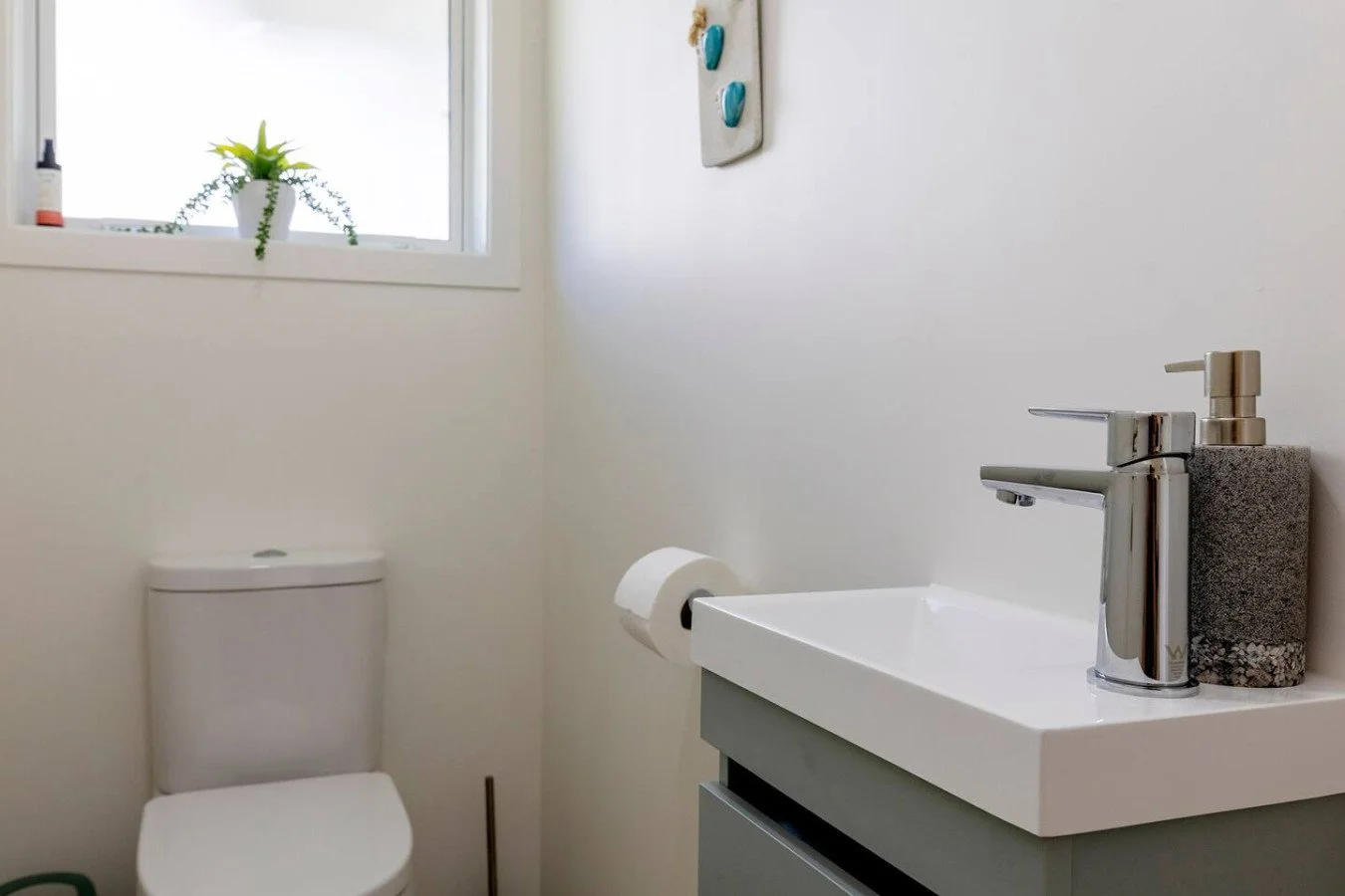 A small bathroom with a white toilet, a new small sink with a chrome faucet, a window with a potted plant on the windowsill, a bottle on the windowsill, a roll of toilet paper on a holder, and a soap dispenser on the sink. The walls are painted white