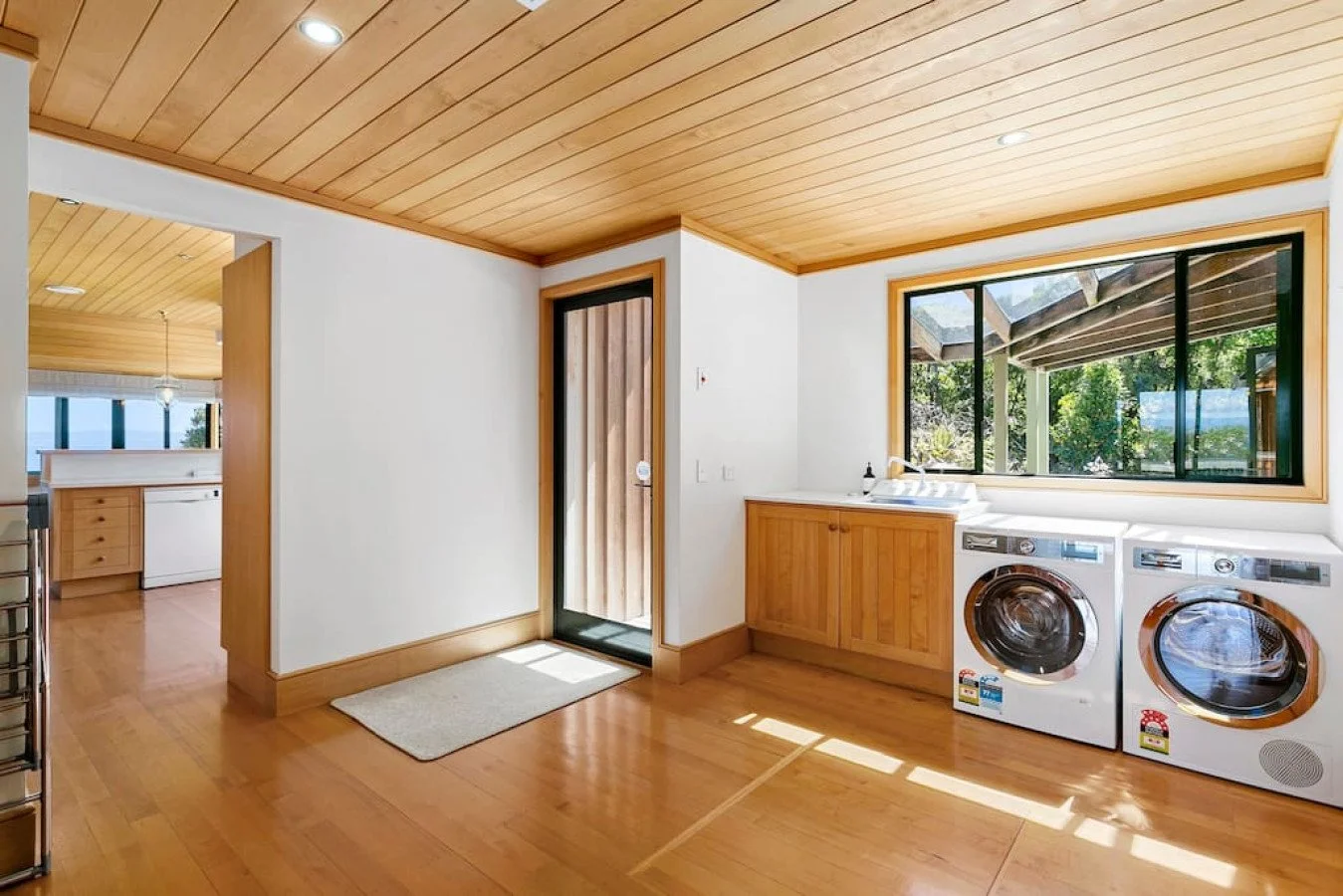 A laundry room with wooden cabinets, a washer and dryer, a large window showing greenery outside, and a door leading outside.