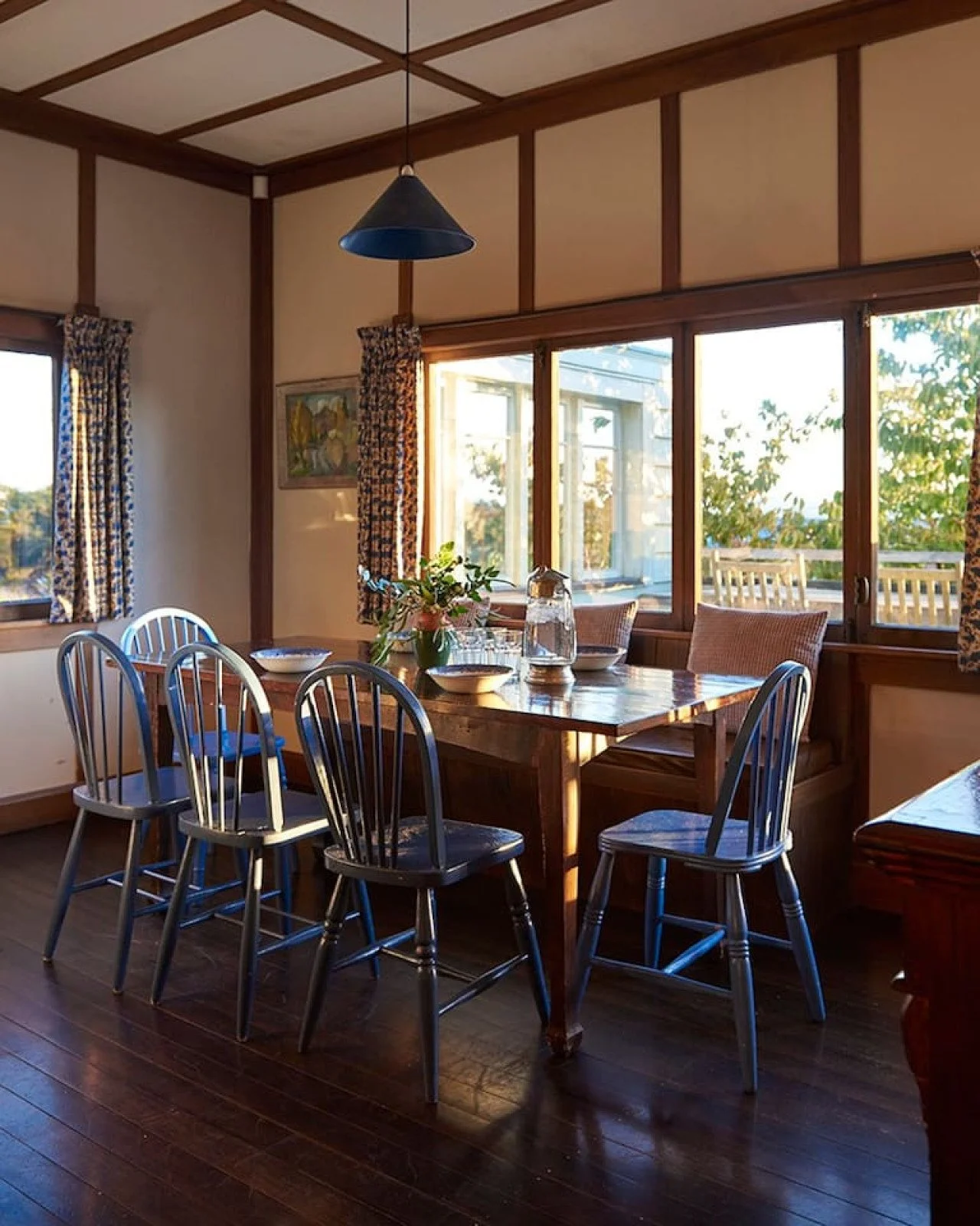 Cozy dining room with wooden table and five blue chairs, floral centerpiece, glass pitcher, bowls, and large windows with patterned curtains letting in natural light.