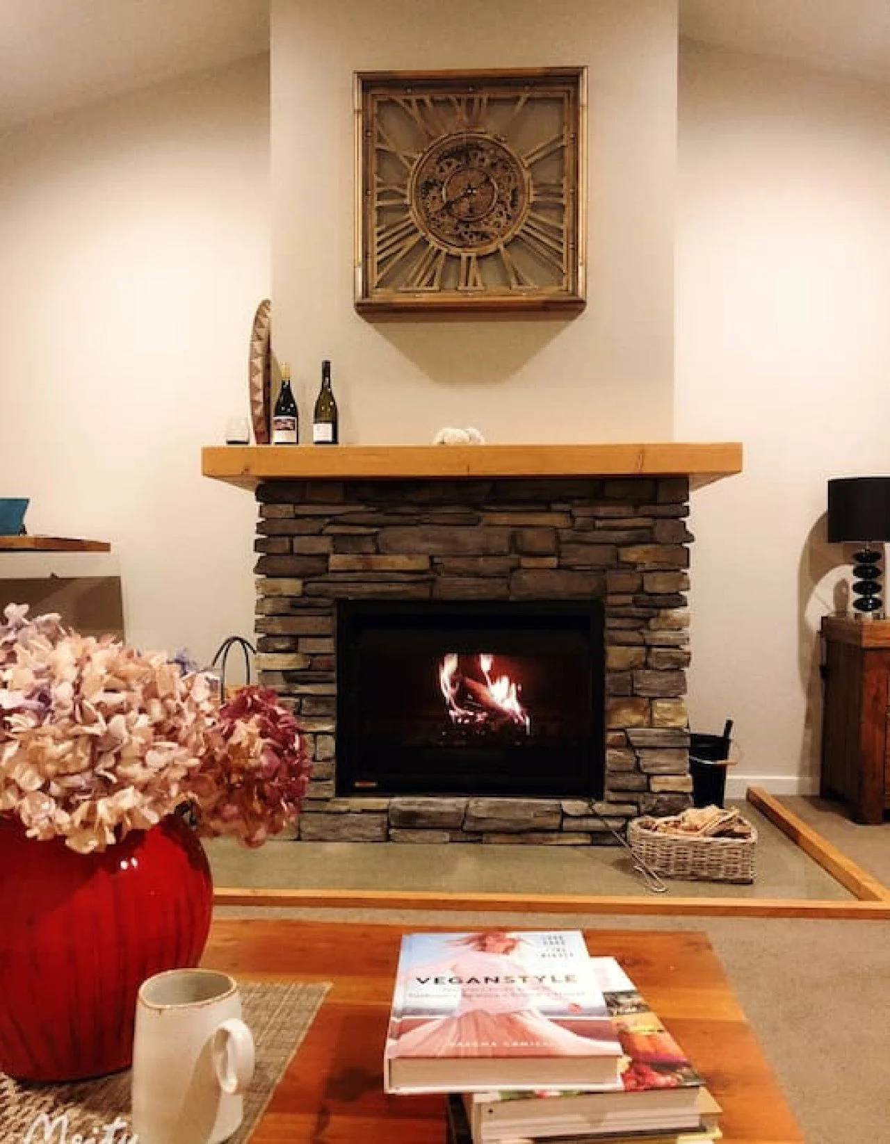 Living room with a stone fireplace, wooden mantel, a large clock above, and a fire burning inside. Decorations include bottles of wine, a vase of pink flowers, books on the coffee table, and a lamp on the side table.