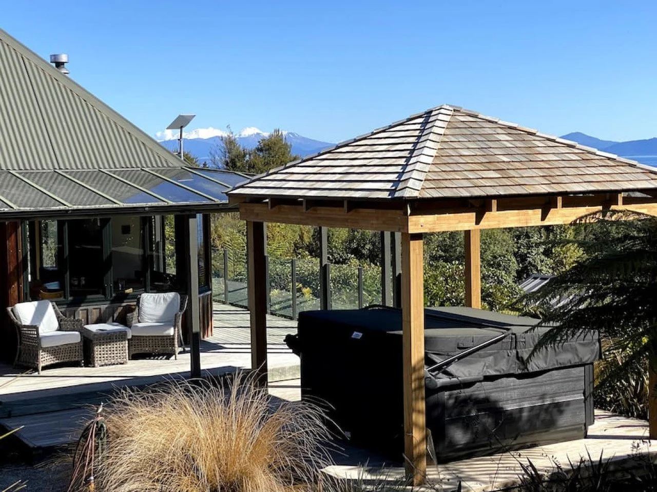 Outdoor deck with seating area, hot tub under a small wooden pavilion, and a house with a metal roof; mountains and trees in the background.