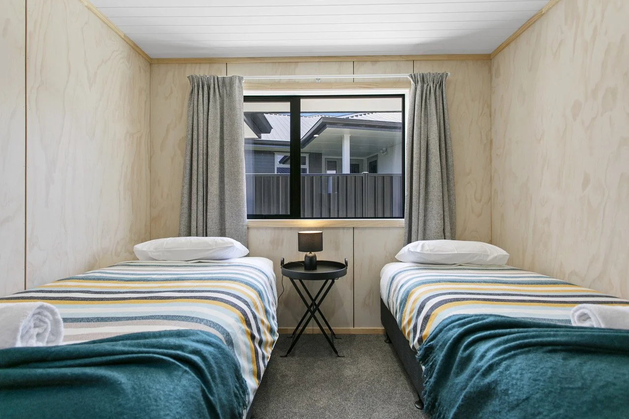 A bedroom with two twin beds with striped bedding, a small black nightstand with a lamp between the beds, and a window showing a neighboring house with a porch.