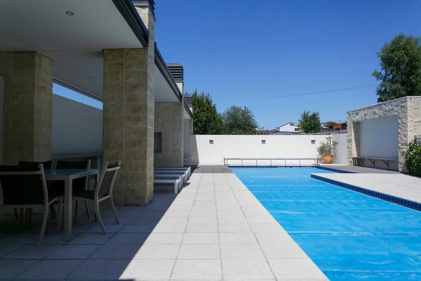 Modern outdoor pool area with patio furniture, stone columns, a white wall with small square lights, and a potted plant against a clear blue sky.
