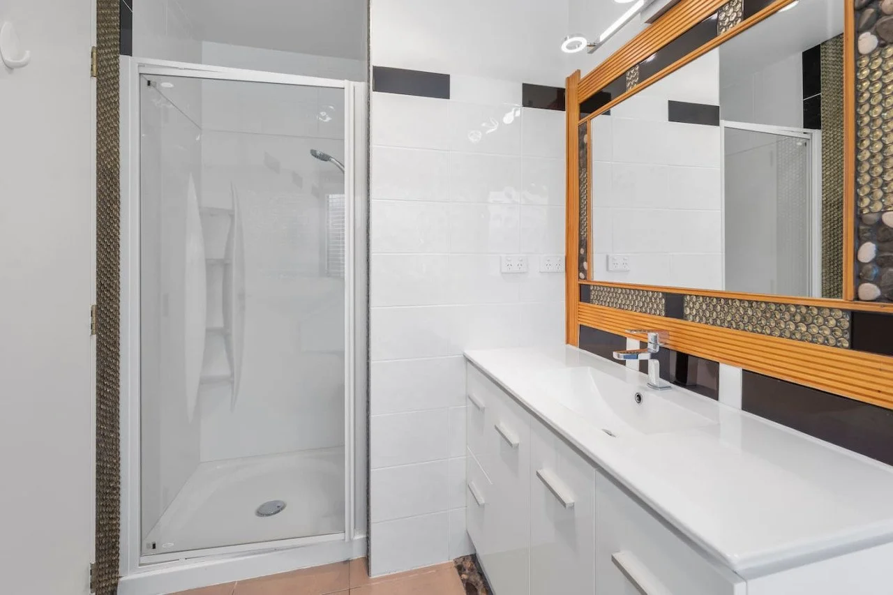 Modern bathroom with a glass shower stall, white vanity with sink, a mirror framed with wood and decorative tiles, and a patterned wall.