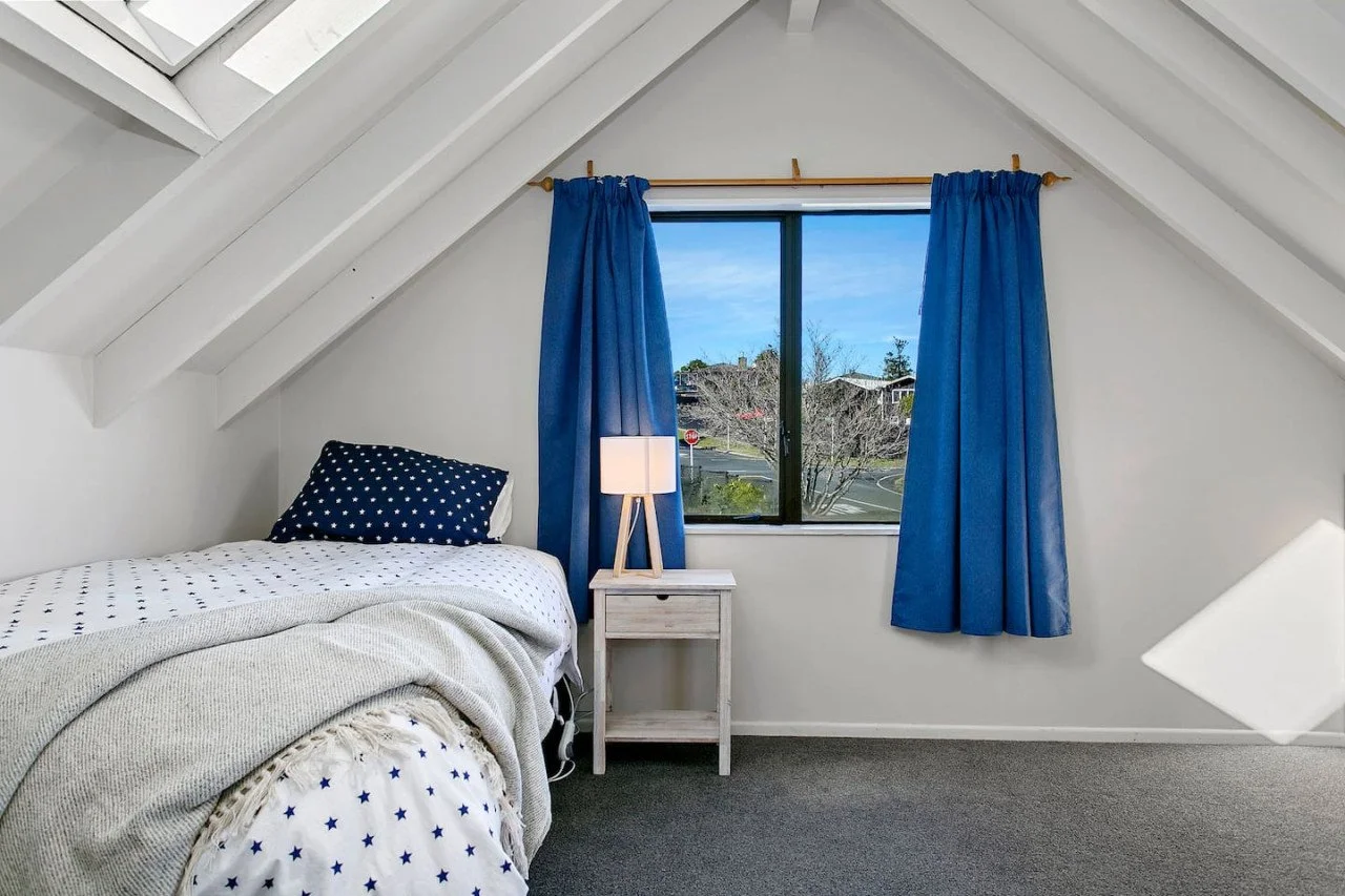 A cozy bedroom with a sloped ceiling, featuring a bed with blue and white star-patterned bedding, a small nightstand with a lamp, and blue curtains on a window showing a street view outside.