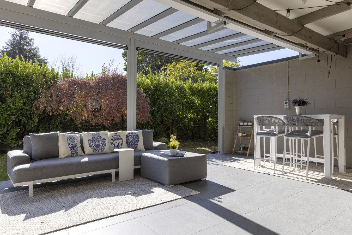 Modern outdoor patio with a gray sofa with decorative pillows, a small side table with a plant, a bar-height table with two chairs, and a garden with trees and bushes in the background.