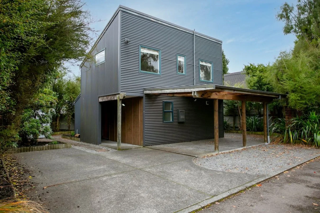 Modern two-story house with black metal siding and a wooden carport on the right side, surrounded by trees and greenery.