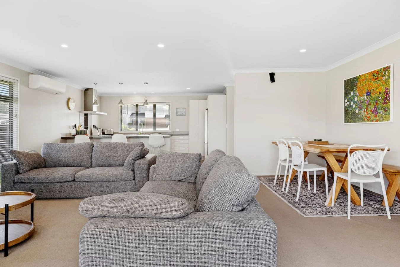 Open-concept living and dining area with gray sofas, a kitchen with white cabinets and bar stools, and a dining table with white chairs, artwork on the wall, and beige carpet.