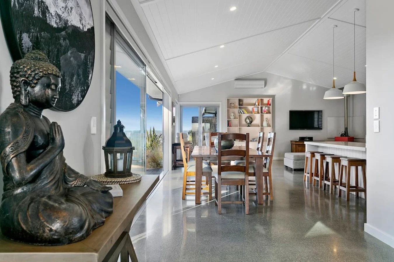 Modern living room with a dining table, bar stools, a TV, and built-in bookshelves. Large windows and sliding glass doors show a view outside.