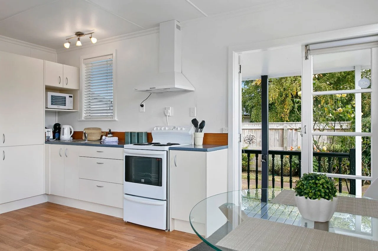 Kitchen with white cabinets, a stove, microwave, kettle, and a view of a backyard with trees and a wooden fence.