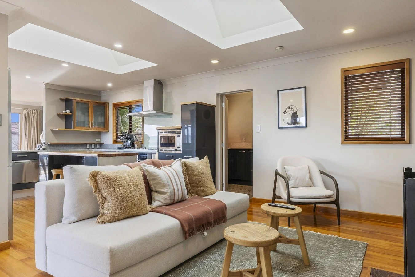 Living room with white sofa, beige and striped pillows, wooden coffee tables, armchair, artwork, windows with wooden blinds, and kitchen area in background