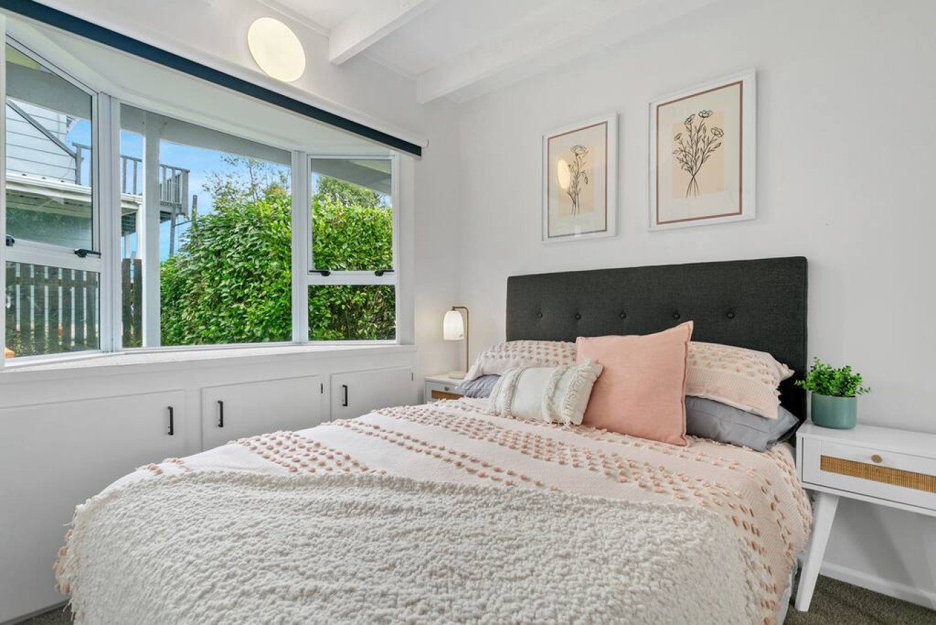 A cozy bedroom with a large window showing greenery outside. The bed has a black tufted headboard and is covered with pink and beige bedding, including multiple pillows. There is a white nightstand with a small plant and a lamp, and two framed floral