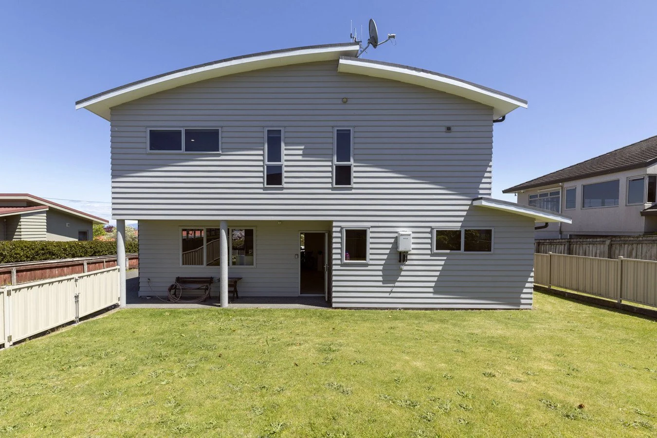 Rear view of a two-story house with light gray siding, a curved roof, and a fenced backyard with green grass, under a clear blue sky.