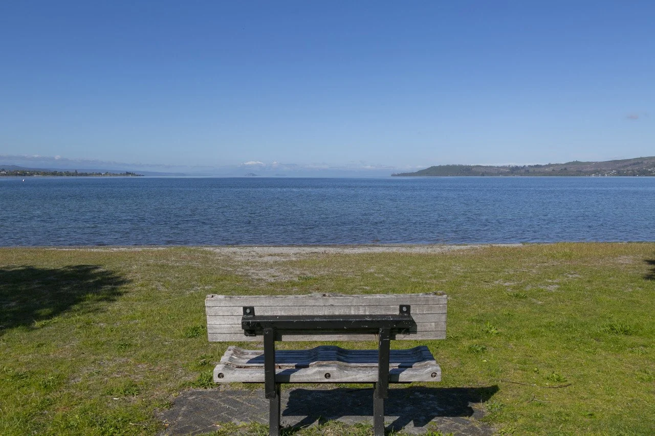 A wooden park bench facing a calm body of water with a distant shoreline and hills under a clear blue sky.