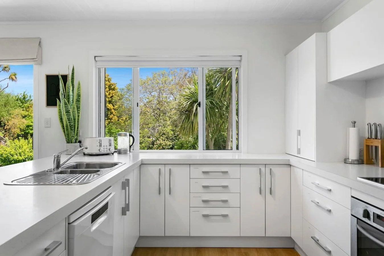 Bright white kitchen with large window overlooking green trees, countertop with toaster and glass pitcher, and various kitchen appliances.