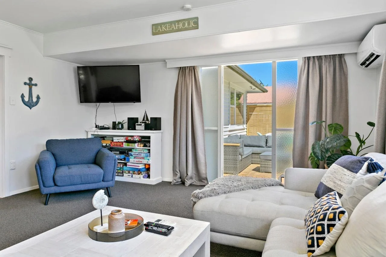Living room with a white couch, blue armchair, and a coffee table, with sliding glass door leading to a patio. There is a wall-mounted TV, a bookshelf filled with board games, and nautical decor.