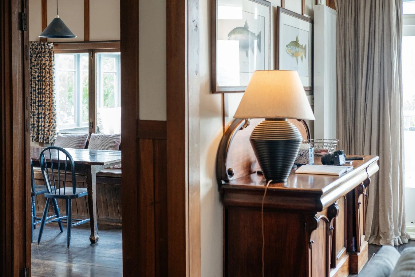 A cozy room with wooden furniture, framed fish artwork, a lamp on a sideboard, and a dining area with a table, blue chair, and window with floral curtains.
