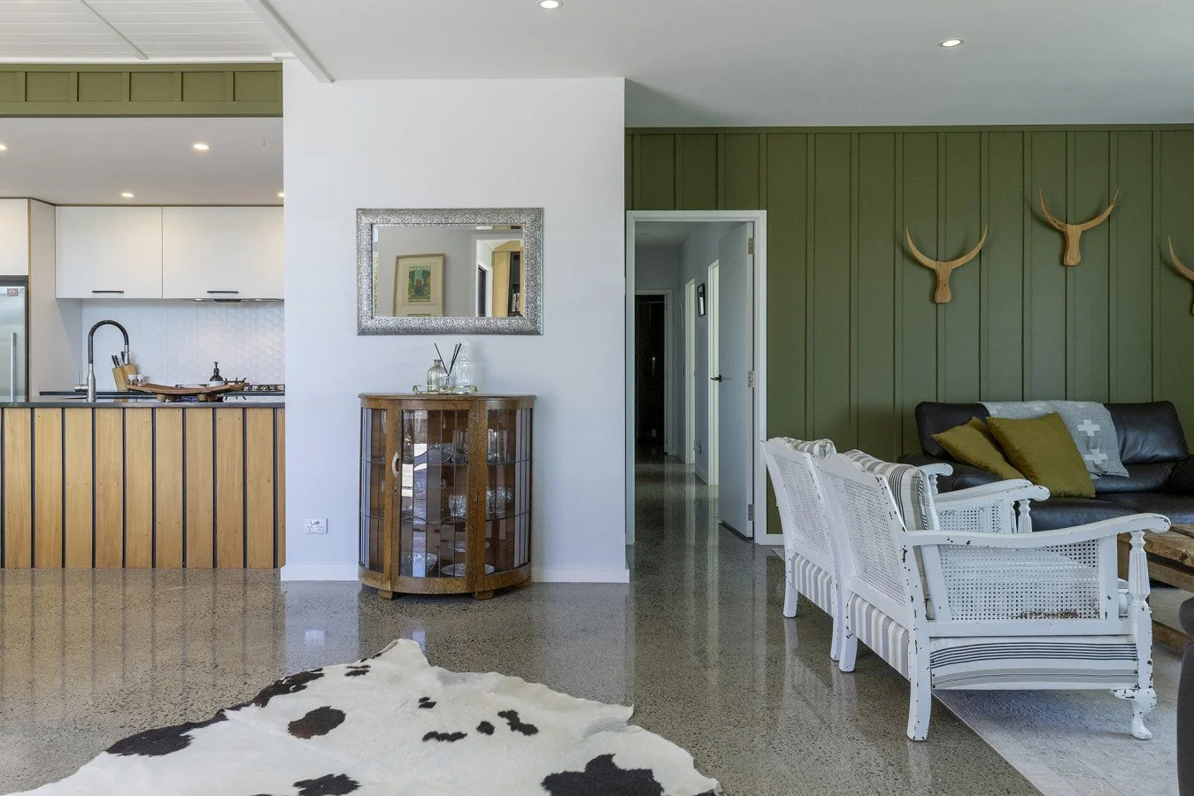 Living room with green wall, white and black chairs, black leather sofa with pillows, cowhide rug, and open kitchen in background with white cabinets and wooden accents.