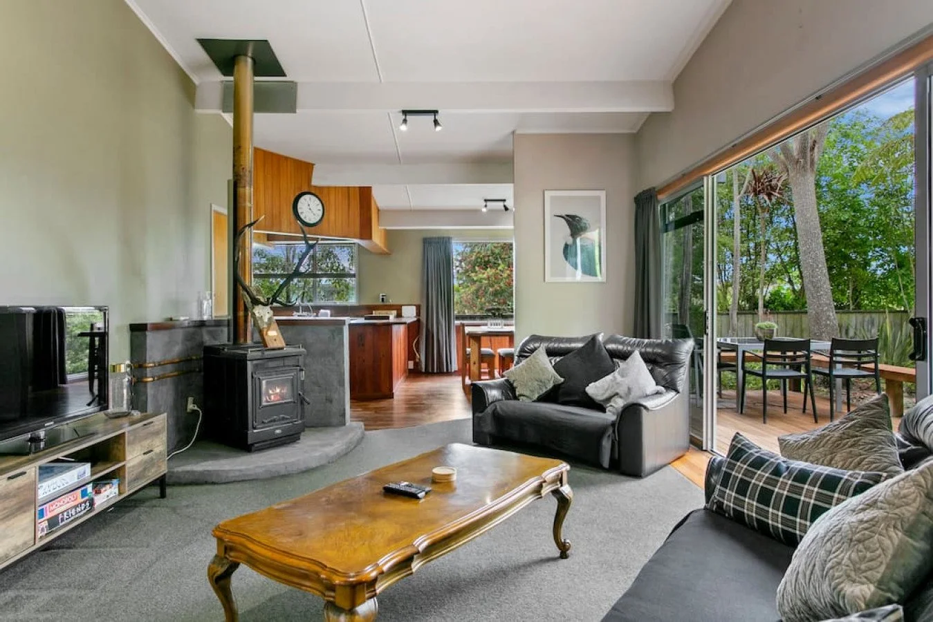 Living room with a wood stove, a black leather sofa, a plaid armchair, and a wooden coffee table. Sliding glass door opens to a deck with outdoor furniture and trees.