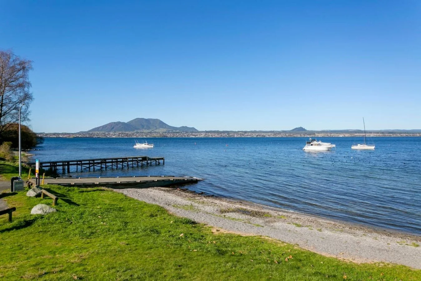 A peaceful lakeside scene with three white boats floating on calm water, a wooden dock extending into the lake, lush green grass and trees along the shore, and mountains in the background under a clear blue sky.