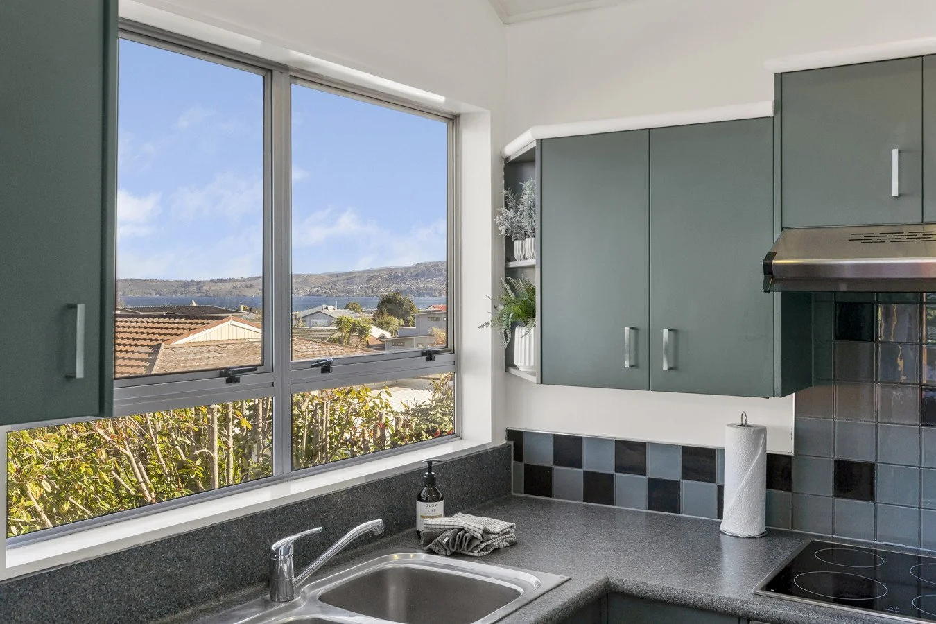 A kitchen with a large window showing a view of rooftops, trees, and distant hills under a partly cloudy sky. The kitchen has gray cabinets, a black tiled backsplash, a gray countertop, a sink with a chrome faucet, a soap dispenser, a paper towel rol