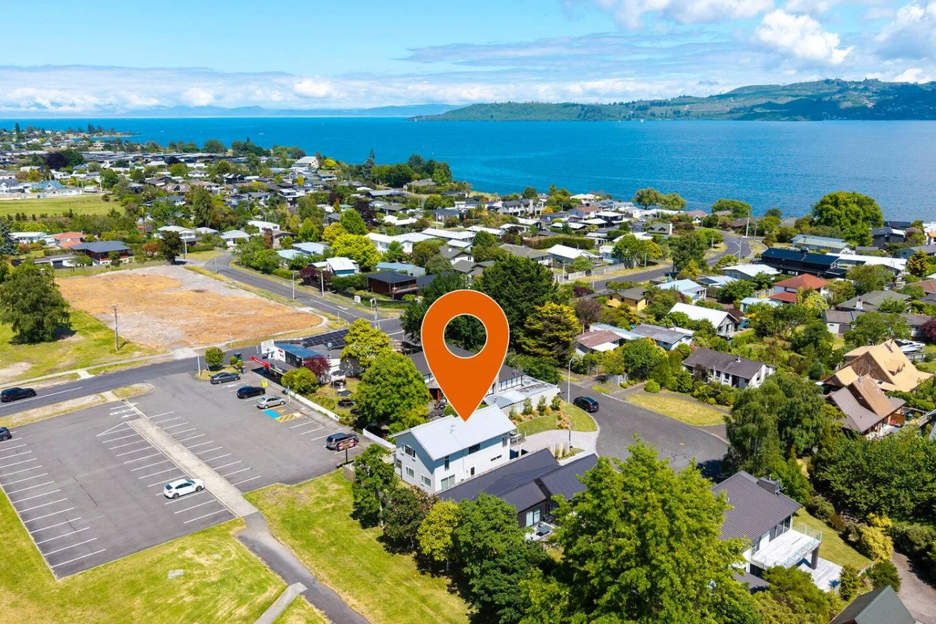 Aerial view of a coastal residential neighborhood with houses, trees, parking lots, and a large body of water in the background.