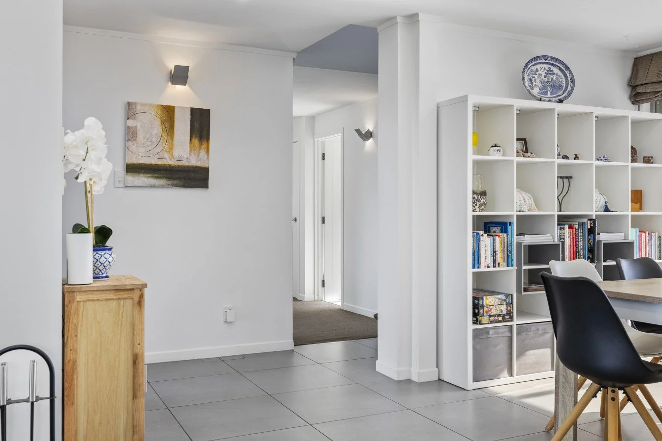 Interior view of a modern home with white walls, tiled floor, a small wooden table with a potted orchid, an abstract painting, and a large white bookshelf filled with books and decorative items. A hallway is visible in the background.