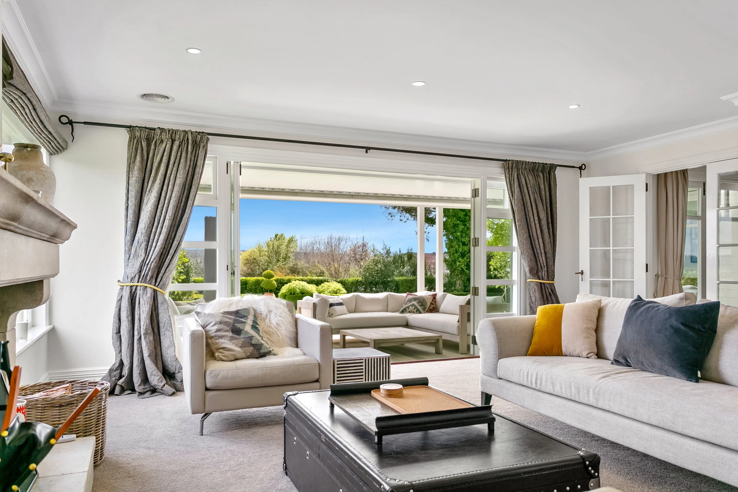 Living room with open sliding glass doors leading to a patio with outdoor seating, beige sofas, colorful pillows, and view of a green garden with trees.