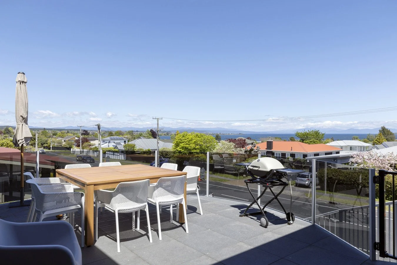Outdoor balcony with a wooden dining table, white chairs, a closed patio umbrella, a barbecue grill, and a scenic view of trees, houses, water, and mountains in the distance under a clear blue sky.