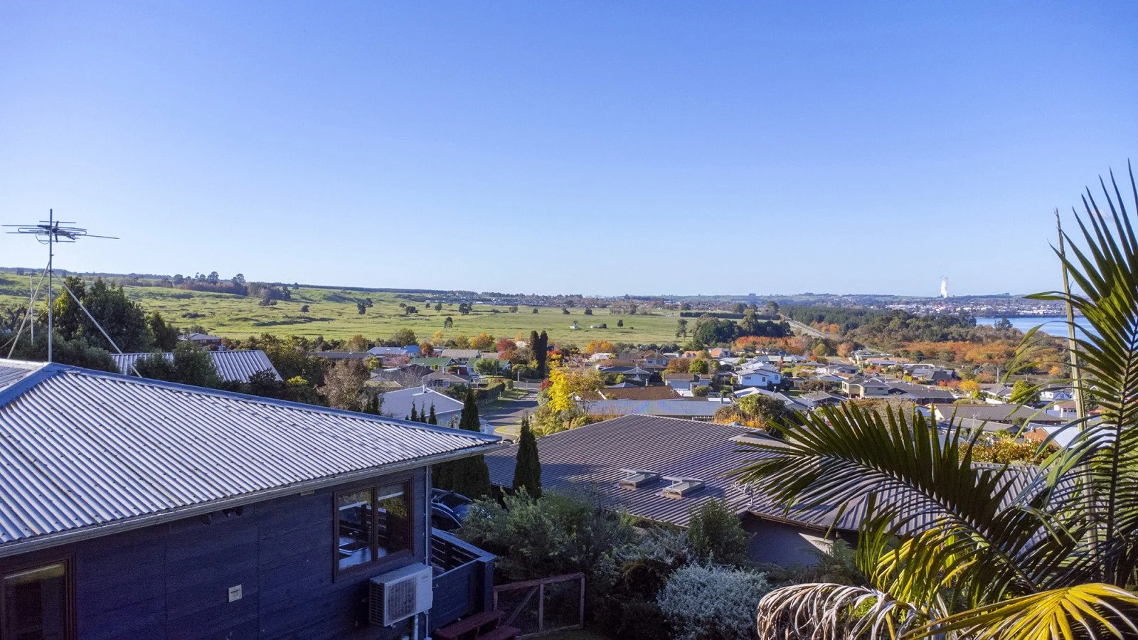 View from a backyard showing rooftops, trees, green fields, and a clear blue sky in the distance.