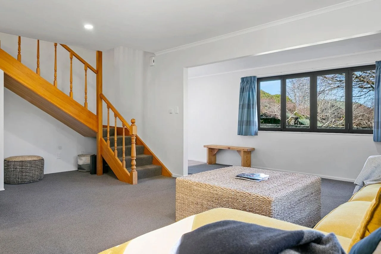 Living room with a staircase to the left, a large window with blue curtains, a yellow sofa, a wicker coffee table with magazines, a wooden bench by the window, and neutral-colored walls and carpet.