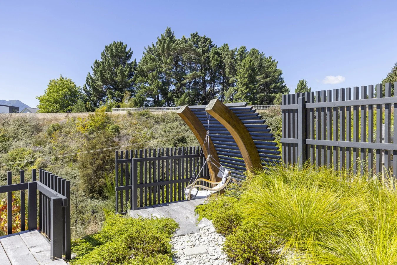 Outdoor patio area with a hanging swing chair beneath a wooden shade structure, surrounded by green bushes and fenced with black railing, with trees and mountains in the background.