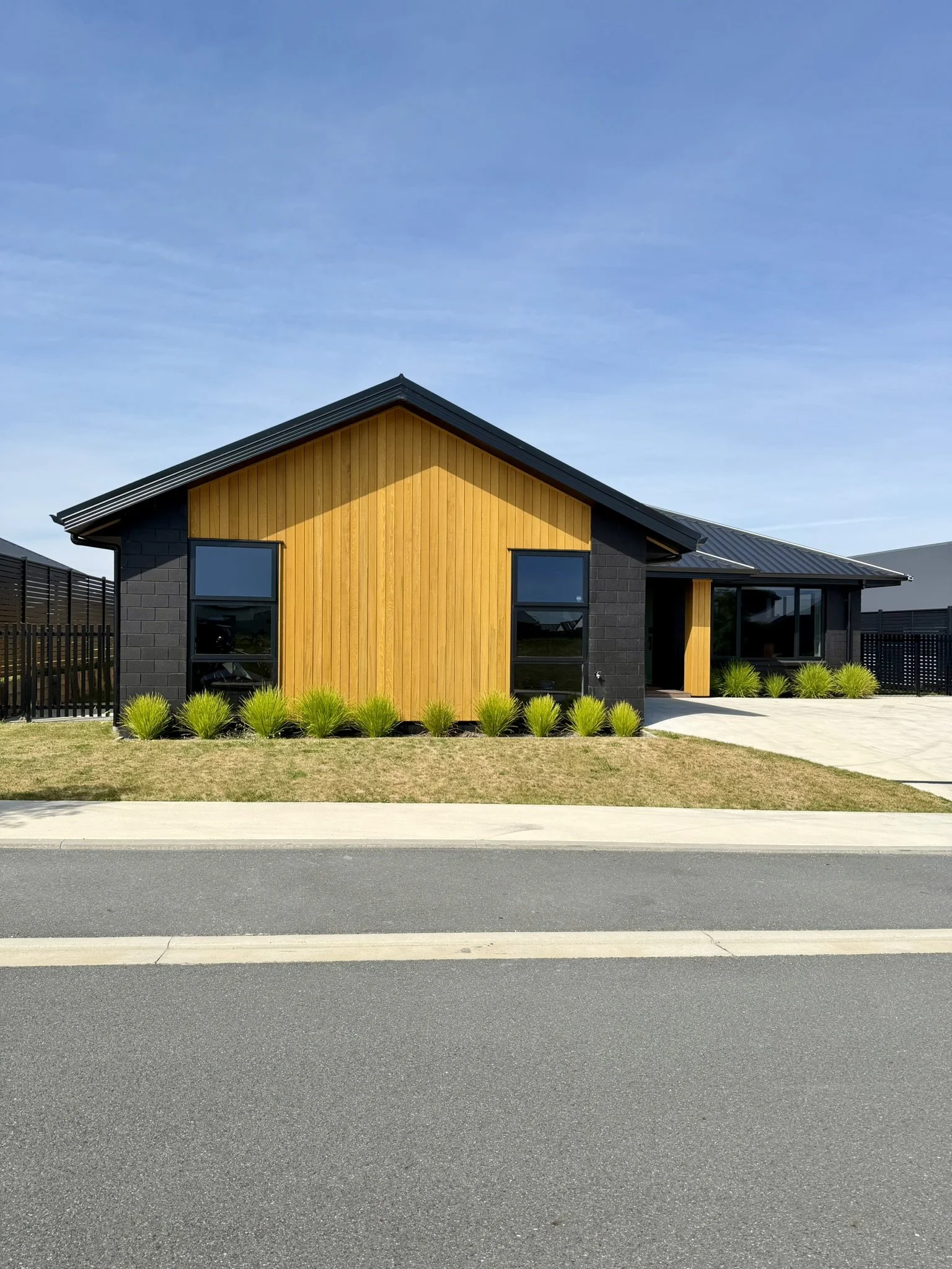 Modern house with black brick and yellow wooden panel exterior, large windows, and a landscaped front yard with small green bushes, situated beside a paved street under a blue sky.