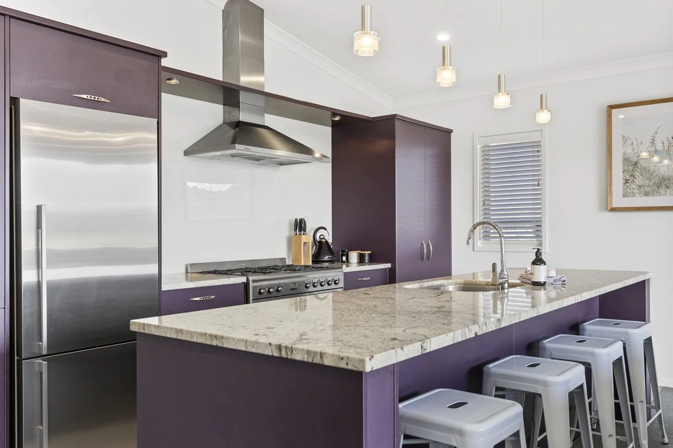 Modern kitchen with purple cabinets, a granite countertop island, stainless steel appliances, and white bar stools, with a window and framed picture on the white wall.