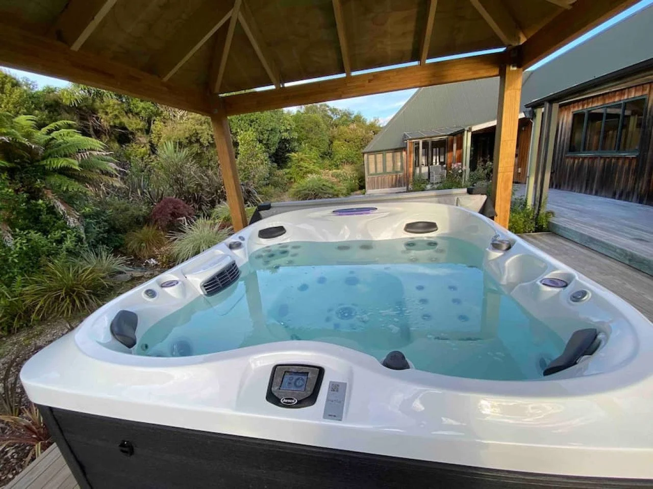 A hot tub filled with water on a deck under a wooden pavilion, surrounded by greenery and outdoor buildings.