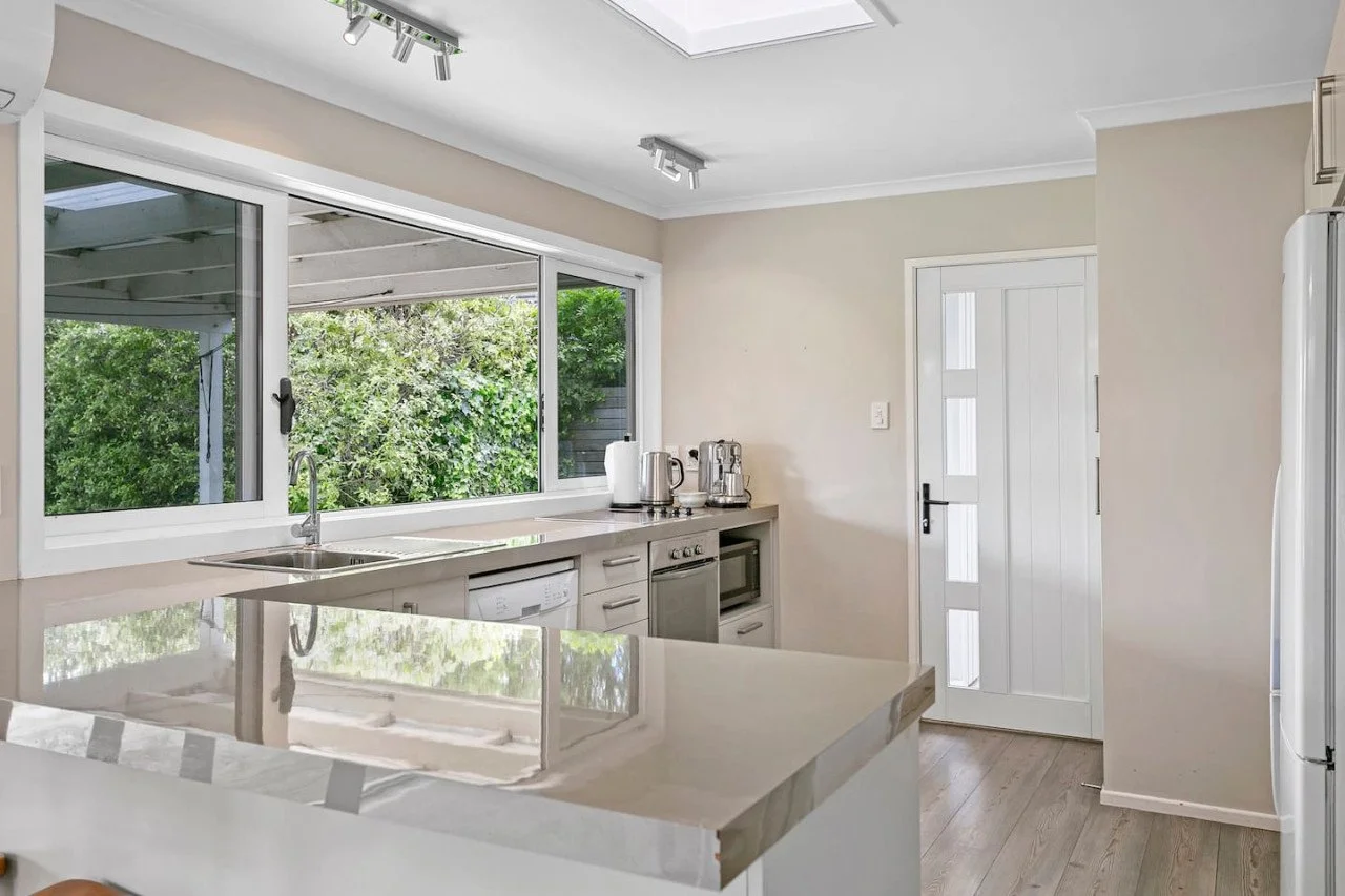 A modern kitchen with a large window overlooking greenery, a white countertop, and built-in appliances including a microwave and dishwasher. There are electric kettles on the counter, and a white door with glass panels leading outside.