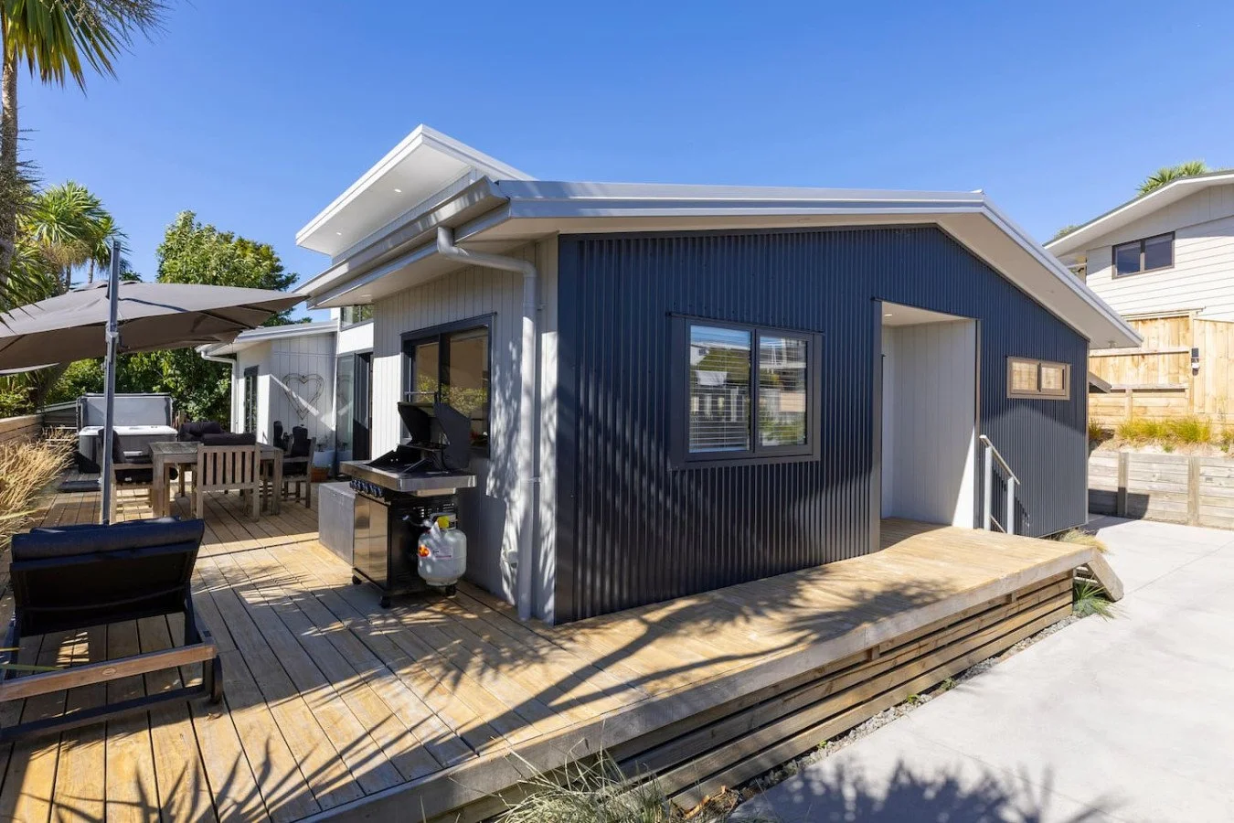 A modern backyard with a wooden deck, outdoor furniture, a grill, and a house with dark and light siding under a sunny sky.