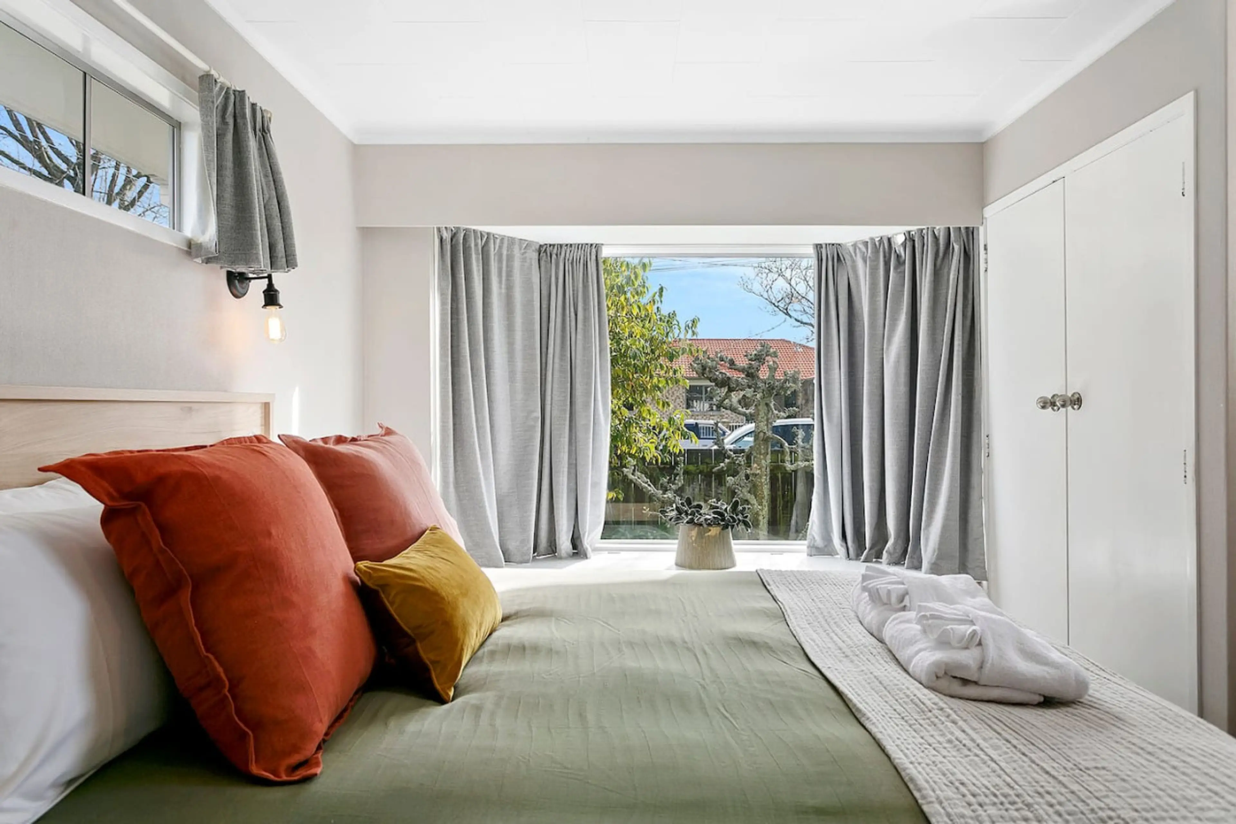 A neatly made bed with orange and mustard pillows, placed in a bright room with large windows and gray curtains, overlooking a garden outside.