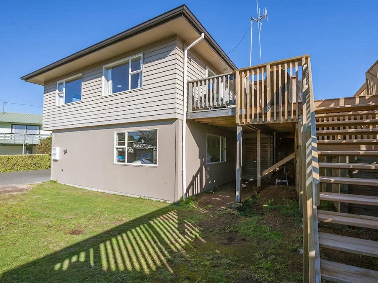 A two-story house with horizontal siding and a deck on the upper level, supported by wooden posts, with staircase leading up to it, and a grassy yard underneath, set against a clear blue sky.